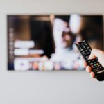 Person using a TV remote control with a blurred television screen in the background.
